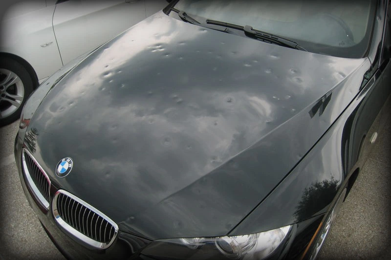 The cloudy sky reflected on the hood of a black car in need of hail repair.