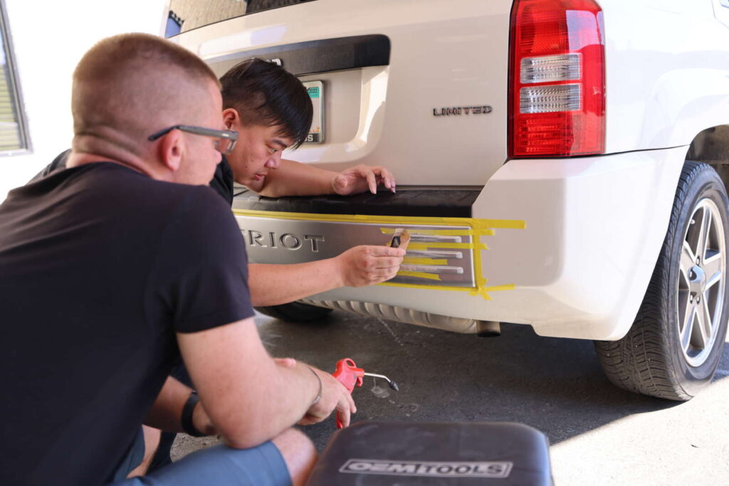 Two men are repairing a scuffed bumper on a white SUV.