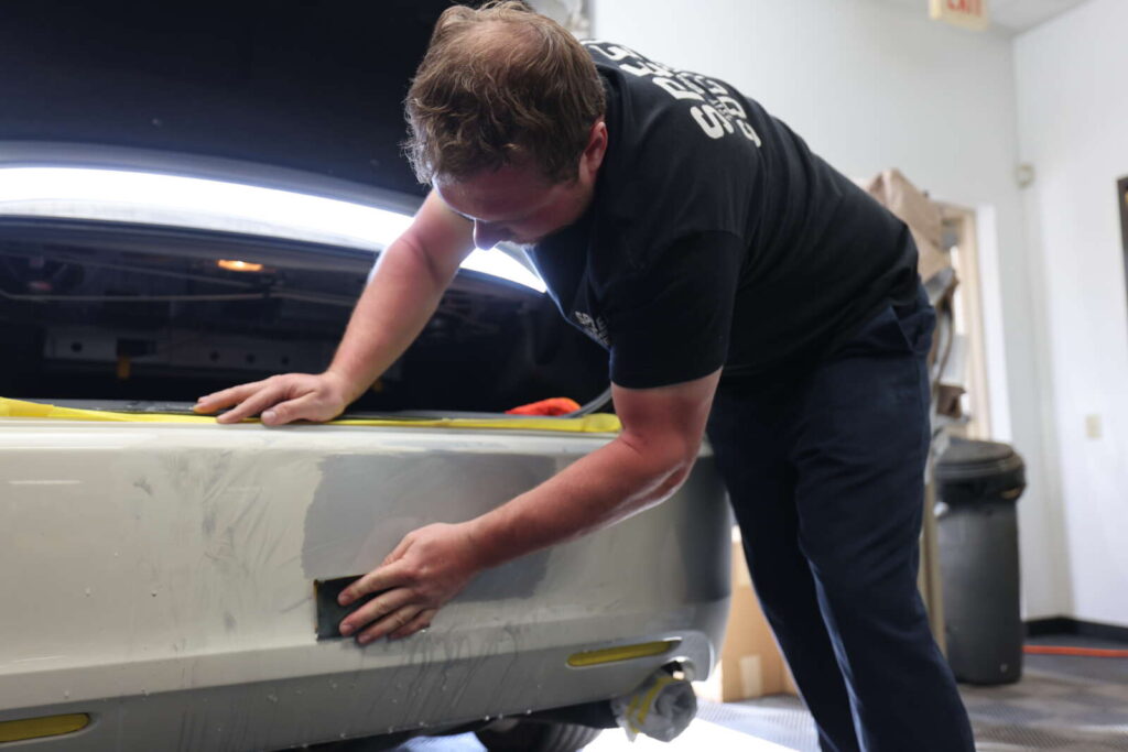 A man is repairing a scuffed bumper on a car in a garage.