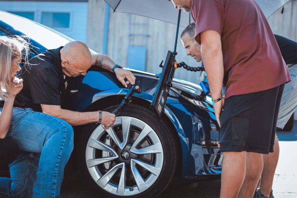 Two men inspect a blue car's front wheel under an umbrella, possibly examining the Paint Protection Film, while a woman and a third man observe.