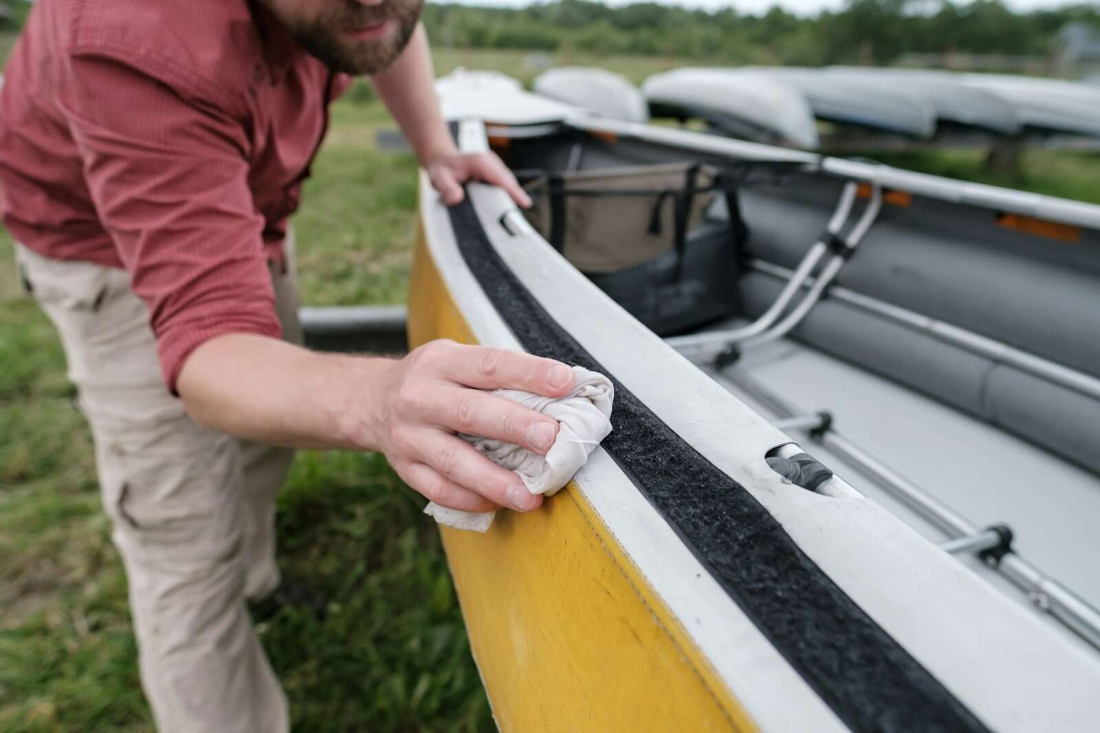 A person in a red shirt and beige pants uses a cloth to clean the edge of a yellow canoe, which is positioned on grass with other canoes in the background, ensuring it’s spotless for the application of Paint Protection Film.